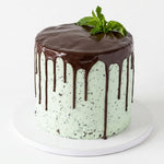 A round chocolate drip cake with mint green frosting speckled with cookie crumbs, topped with a glossy chocolate glaze and a fresh mint leaf, displayed on a white cake stand against a white background. Choc Mint cake with rich chocolate sponge layers, fresh cream and refreshing mint chocolate filling, beautifully decorated on a white cake board by Cake Gurus in Ruaka, Nairobi.