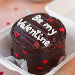 A chocolate bento cake with the phrase 'Be my Valentine' written on top, decorated with red heart shapes and situated inside a white box.