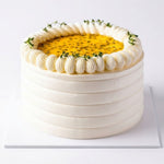 A round white frosted cake with piped cream borders and a yellow passion fruit center, garnished with small green herb sprigs, shown on a white square board against a white background. Passion cake with moist sponge layers, fresh cream and tropical passion fruit filling, decorated on a white cake board by Cake Gurus in Ruaka, Nairobi.