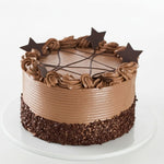 A round chocolate cake with smooth milk-chocolate frosting, piped swirls around the top, chocolate drizzle on top, and chocolate star decorations, sitting on a white plate against a white background. Tiramisu cake with layers of soft coffee soaked sponge, fresh cream and rich mascarpone style filling, dusted with cocoa and photographed on a white cake board by Cake Gurus in Ruaka, Nairobi.