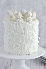 A white coconut cake with coconut flakes on the sides and topped with coconut cream and cherries as a sweet treat by Cake Gurus Ruaka Nairobi with deliveries in Nairobi and its environs.