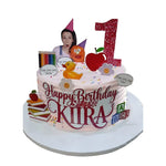 A custom round birthday cake with pink frosting, decorated with a large red glitter number 1 topper, a photo of a woman, a yellow rubber duck, a red apple, flowers, books, and colorful alphabet tiles. The cake is displayed on a white round cake board against a white background. **Miss Racheal themed 1kg vanilla birthday cake with custom toppers, custom cakes in Ruaka Nairobi by Cake Gurus**
