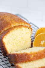A loaf of orange cake with a few slices cut out, displayed alongside fresh orange slices.