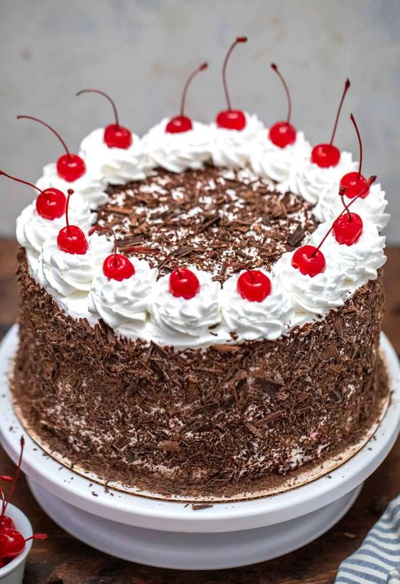 A Black forest cake with white whipped cream and cherries on top, garnished around the edge with chocolate shavings,  as a sweet treat by Cake Gurus Ruaka Nairobi with deliveries in Nairobi and its environs.