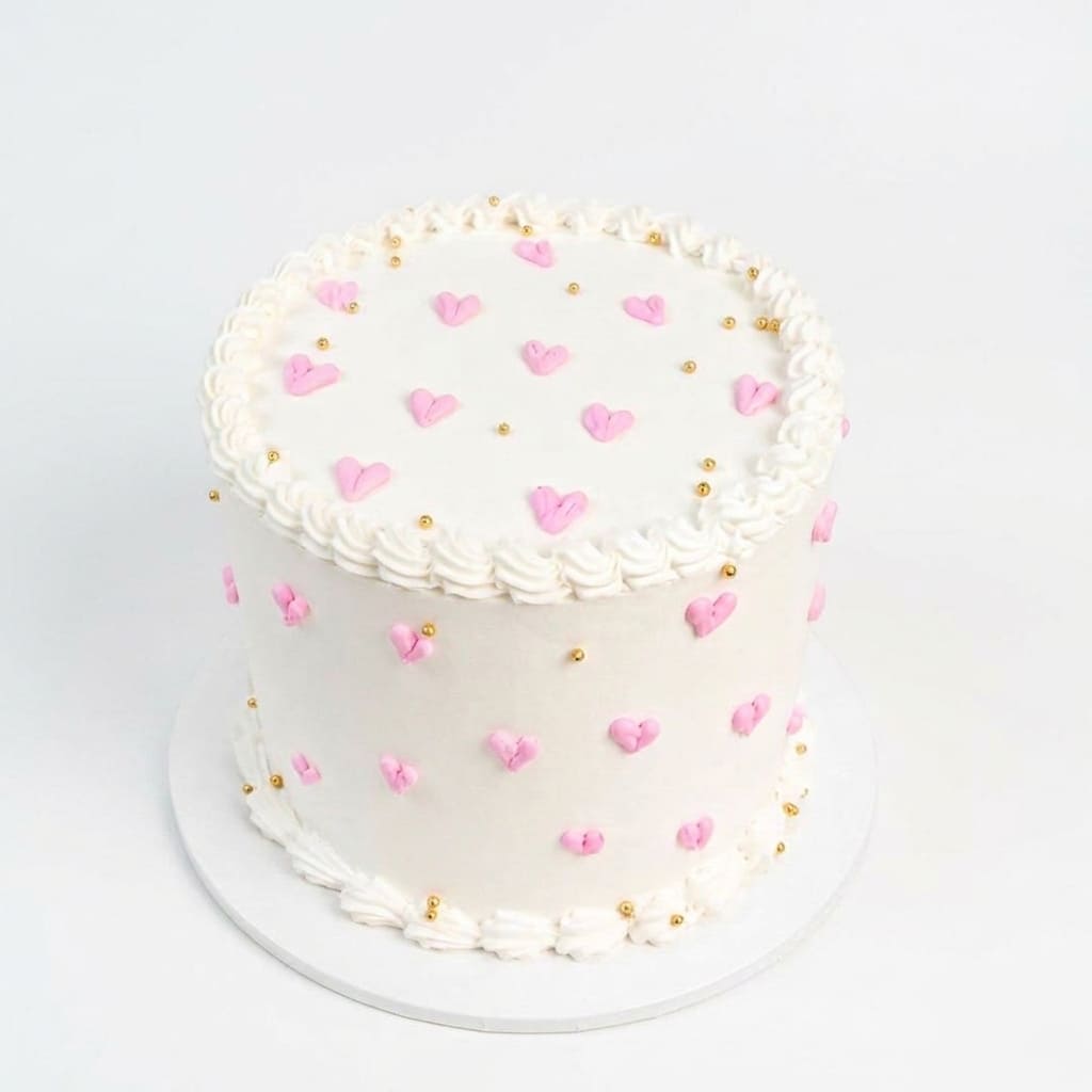 A round white frosted cake decorated with pink heart shapes and small gold dots, presented on a white round board against a plain white background. birthday celebrations, same day cake delivery in Ruaka Nairobi by Cake Gurus