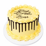 A round birthday cake with pale yellow frosting, piped rosettes around the top and sides, dark chocolate drip decorations, and the message 'Happy Birthday to me, ABE.' written in black icing on top, shown on a white plate against a white background. 0.5kg decorated carrot orange cake from Cake Gurus Ruaka