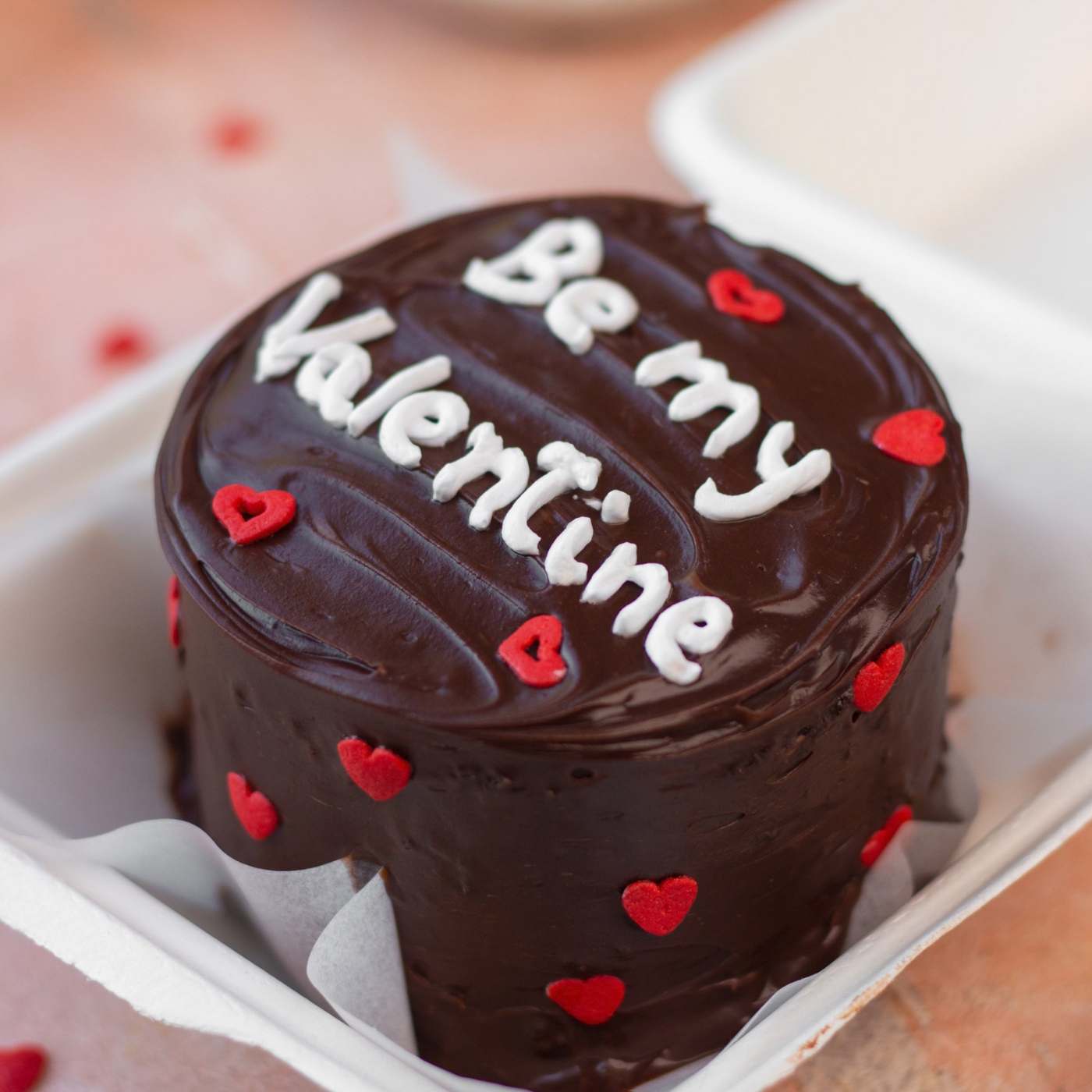 A chocolate bento cake with the phrase 'Be my Valentine' written on top, decorated with red heart shapes and situated inside a white box.