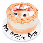 A round birthday cake with a white and peach-colored frosting base, decorated with piped peach rosettes around the top edge, a pink bow, and a realistic bunny face. The cake sits on a white cake board with 'Happy Birthday Bunny' written in black icing. 1kg decorated passion forest cake from Cake Gurus Ruaka