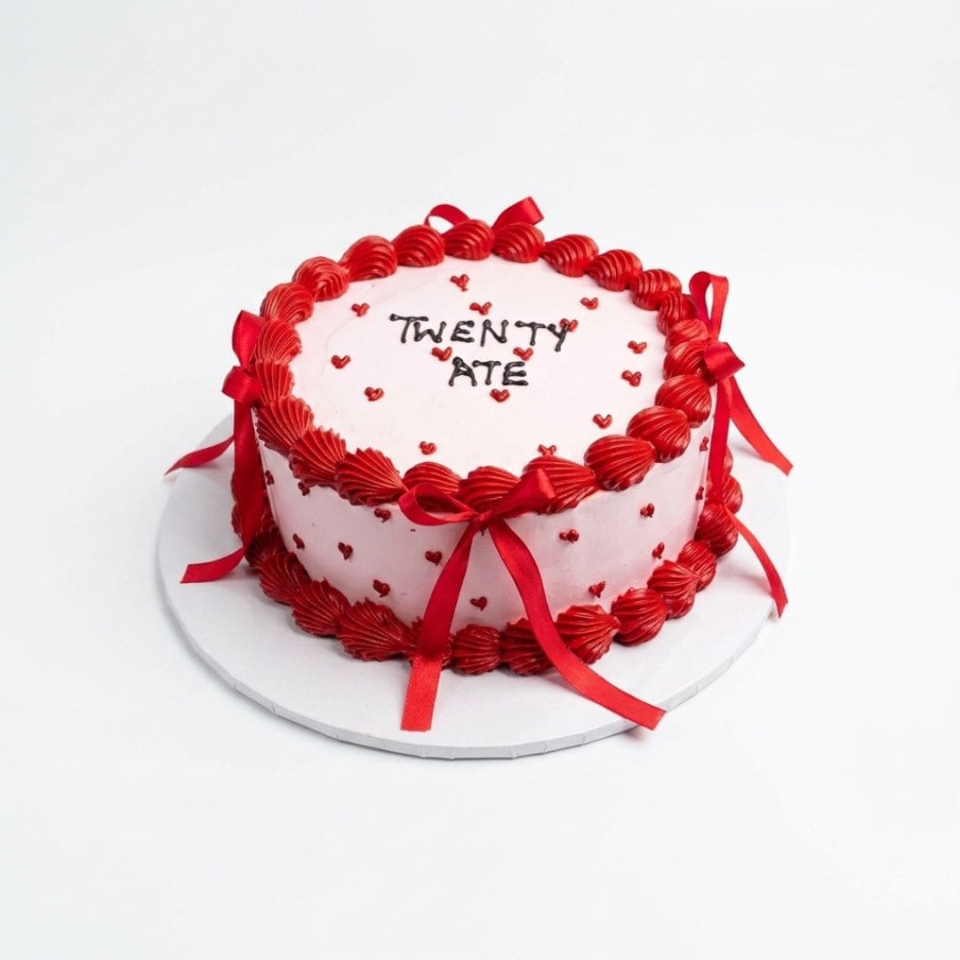 A round birthday cake with white frosting, decorated with red piped borders, red ribbon bows, and small red hearts. The top is written 'TWENTY ATE' in black icing. 1 KG red velvet cake with elegant ribbon decoration, birthday cake from Cake Gurus in Ruaka Nairobi with same day cake delivery