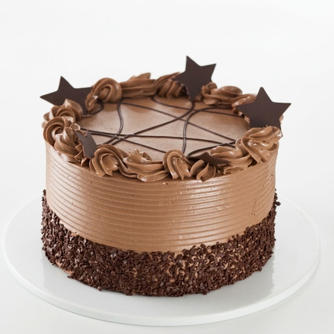 A round chocolate cake with smooth milk-chocolate frosting, piped swirls around the top, chocolate drizzle on top, and chocolate star decorations, sitting on a white plate against a white background. Tiramisu cake with layers of soft coffee soaked sponge, fresh cream and rich mascarpone style filling, dusted with cocoa and photographed on a white cake board by Cake Gurus in Ruaka, Nairobi.