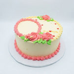 A round frosted cake with a pale cream base, decorated with pink piped roses, green leaves, small red heart details, and a pink rosette border on top and around the base, shown on a white cake board against a light gray background. Banana Birthday Cake for celebrations in Nairobi, freshly baked by Cake Gurus with same day delivery