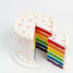 A white frosted rainbow layer cake with a slice cut out and a separate slice beside it, decorated with pink heart sprinkles and gold dots on a white background. birthday celebrations, same day cake delivery in Ruaka Nairobi by Cake Gurus